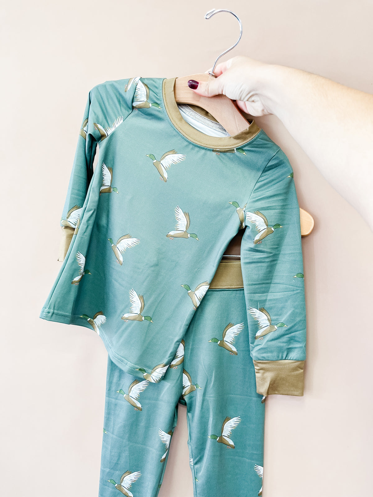 A hand holds up a hanger with the Tiny Tot Bundles Evergreen Mallard Jammies Set, a matching green children’s pajama set featuring a white duck print and beige trim, against a plain light background.
