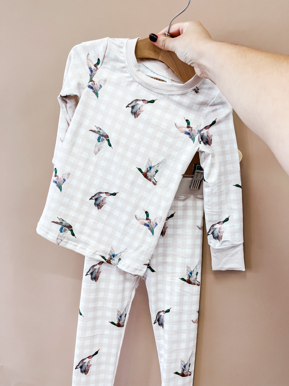 A hand displays the “Morning Flight Jammies Set” by Tiny Tot Bundles, featuring a light checkered pattern with flying duck illustrations, against a plain beige background.