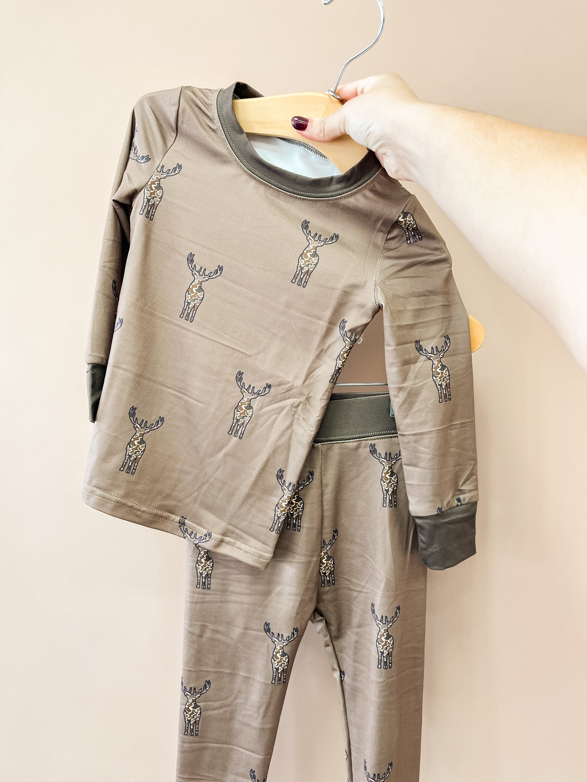 A hand holds a hanger with the Wild Woods Jammies Set by Tiny Tot Bundles, a matching set of brown children’s pajamas adorned with a repeating moose pattern, displayed against a neutral background.
