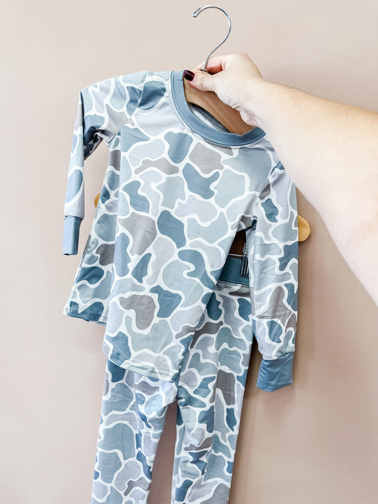 A hand holds up a hanger displaying the Tiny Tot Bundles Wild Country Jammies Set—blue and gray camouflage-patterned children’s pajamas, featuring a long-sleeve top and pants—against a plain beige background.