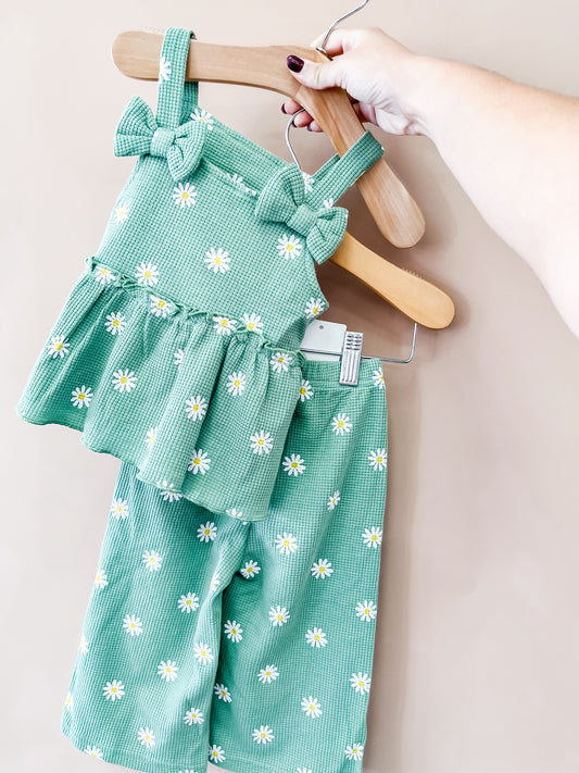 A hand holds a hanger displaying the Tiny Tot Bundles Sage Meadow Pant Set—a toddler outfit in green adorned with white daisy prints. The set features a ruffled top with bow straps and coordinating pants, shown against a neutral background.