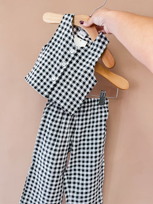 A hand holds the Tiny Tot Bundles Autumn Gingham Pant Set, featuring a black and white checkered sleeveless top and matching pants, set against a plain light brown background.