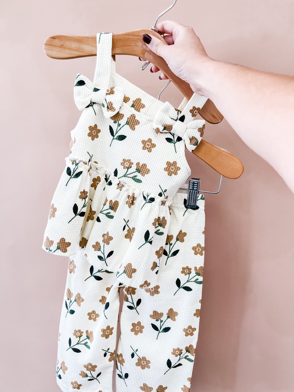 A hand holds a hanger displaying the Tiny Tot Bundles Autumn Wildflower Pant Set, a sleeveless floral-patterned children’s outfit with ruffles on the top and matching pants, set against a plain light pink background.