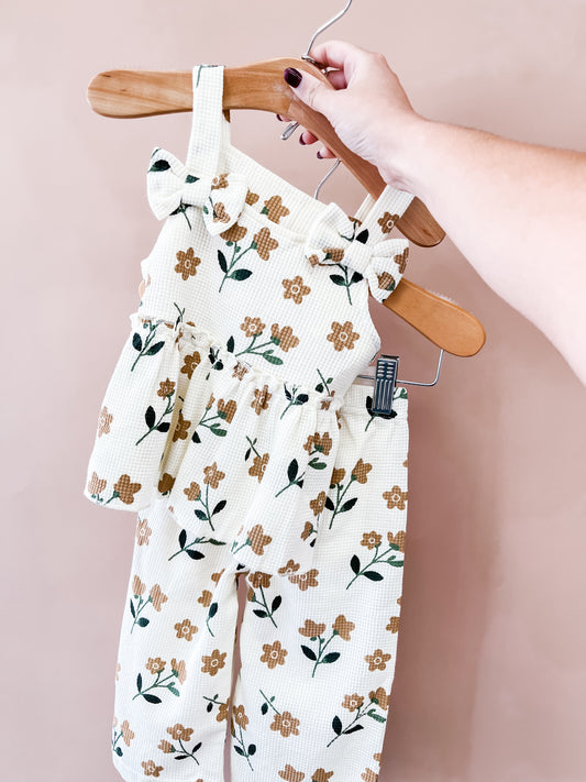A hand holds a hanger displaying the Tiny Tot Bundles Autumn Wildflower Pant Set, a sleeveless floral-patterned children’s outfit with ruffles on the top and matching pants, set against a plain light pink background.