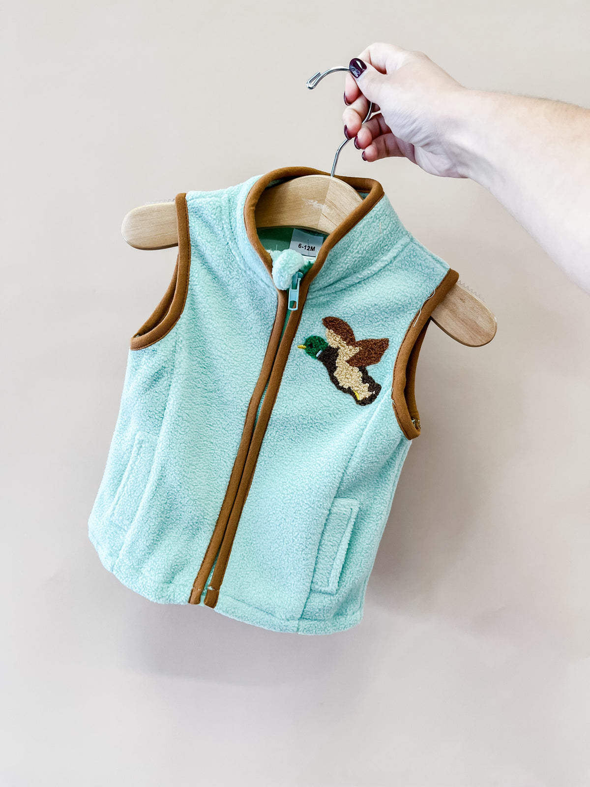 A hand holds a hanger displaying the Tiny Tot Bundles Little Mallard Vest—a light blue fleece vest with brown trim and a duck patch on the chest—set against a plain beige background.