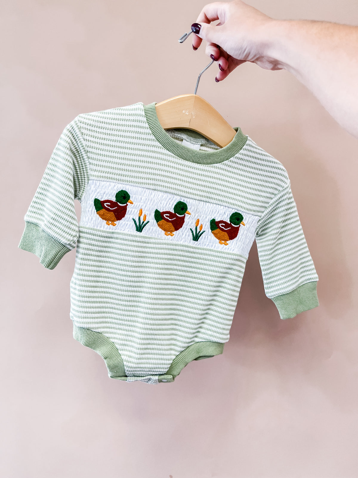 A hand holds the Tiny Tot Bundles Mallard Marsh Long Sleeve Bubble on a hanger, showcasing its green-striped pattern and embroidered trio of ducks with grass on the front.