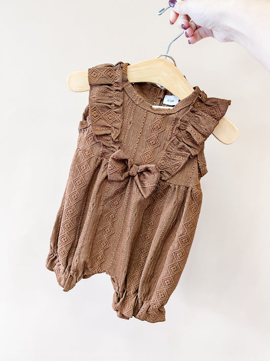 A hand holds a wooden hanger displaying the “Chestnut Ruffle Romper” by Tiny Tot Bundles—a brown, textured baby romper featuring ruffled sleeves, a bow on the chest, and a subtle diamond pattern.