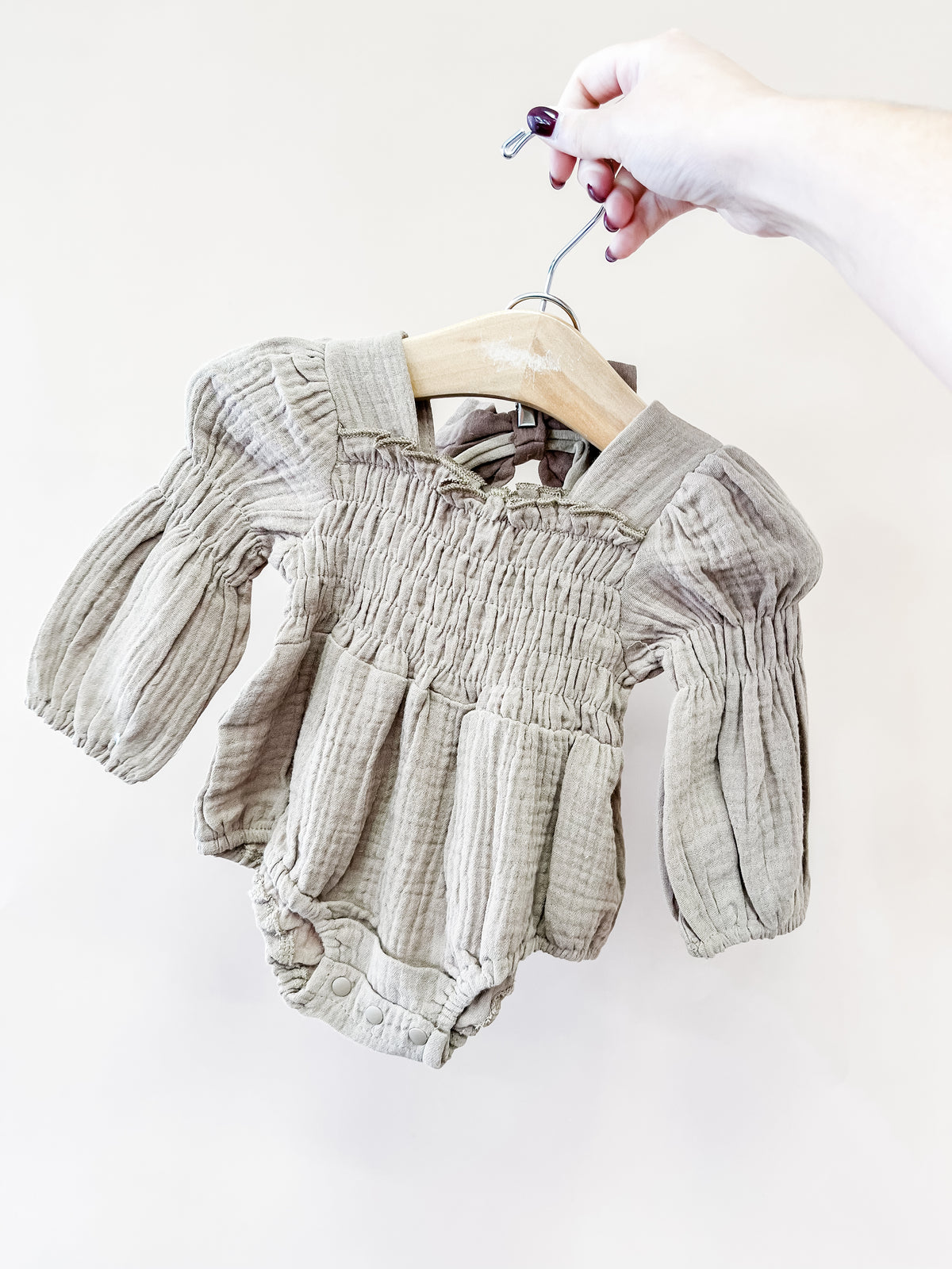A hand holds a wooden hanger displaying the Tiny Tot Bundles Oatmeal Smocked Romper, featuring long sleeves, a smocked bodice, and puffed sleeves against a plain light background.