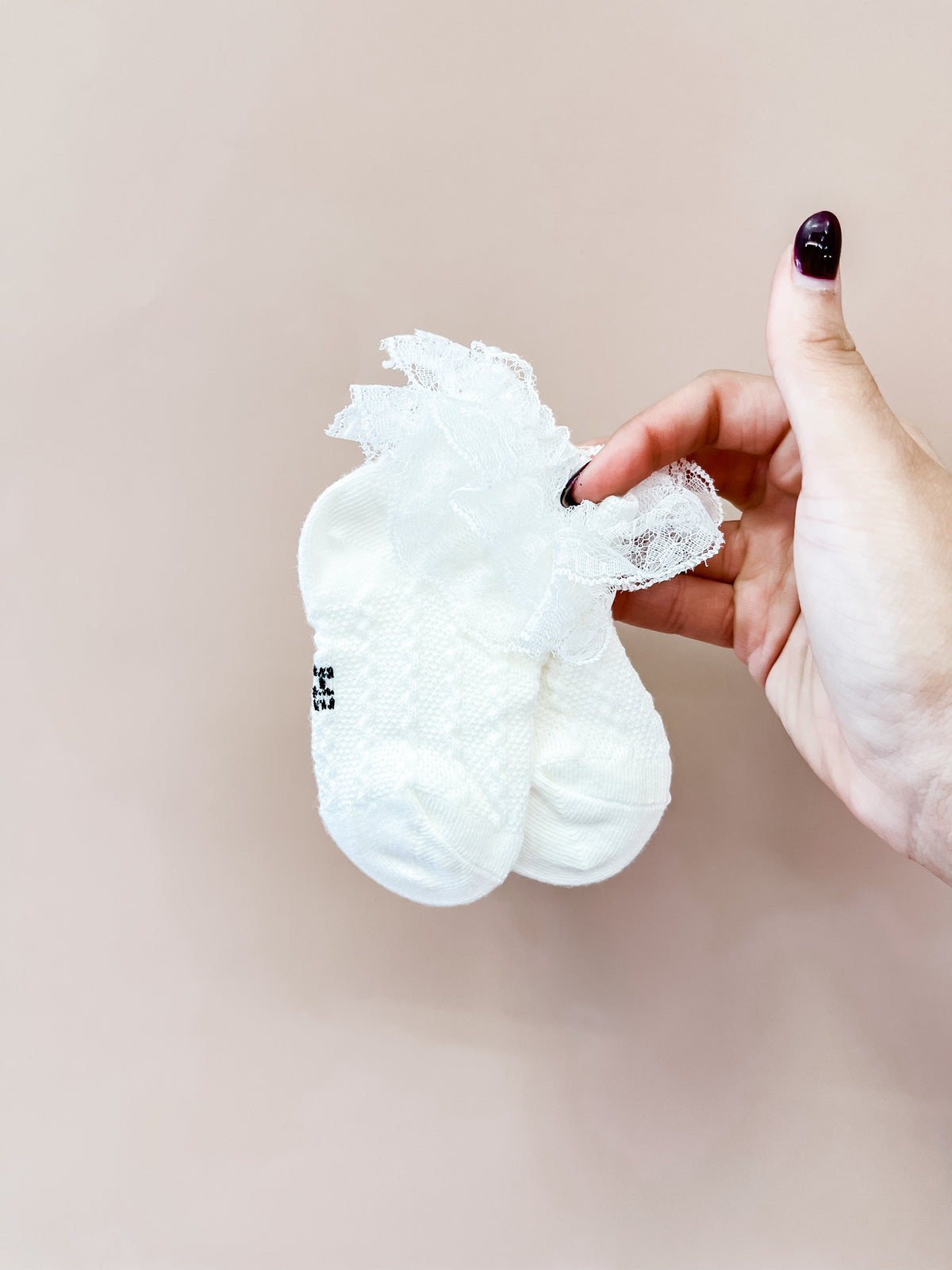 A hand with dark nail polish holds a pair of Tiny Tot Bundles’ Vintage Lace Ruffle Socks against a plain, light beige background.
