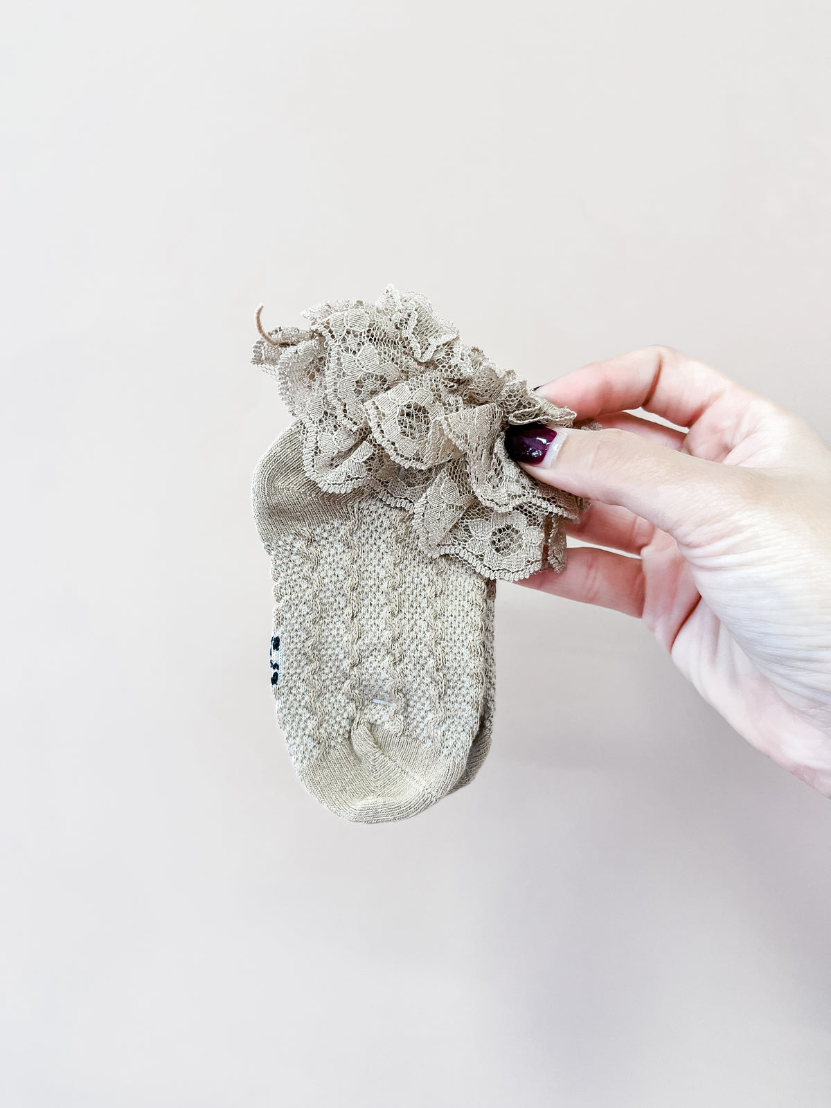 A hand with dark-red nail polish holds up a small beige sock from Tiny Tot Bundles, showcasing their Vintage Lace Ruffle Socks with decorative lace ruffles at the cuff, set against a plain light beige background.