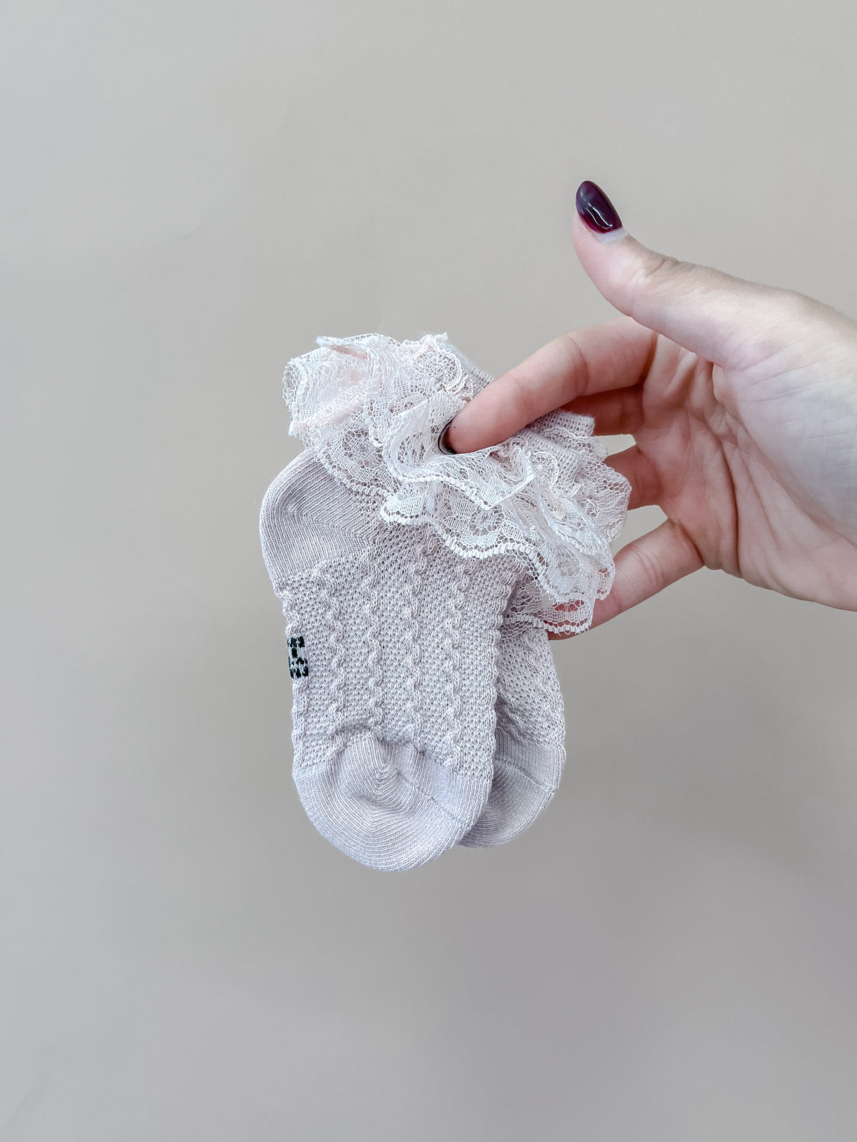 A hand with dark nail polish holds a small pair of Tiny Tot Bundles Vintage Lace Ruffle Socks in pale pink against a plain beige background.