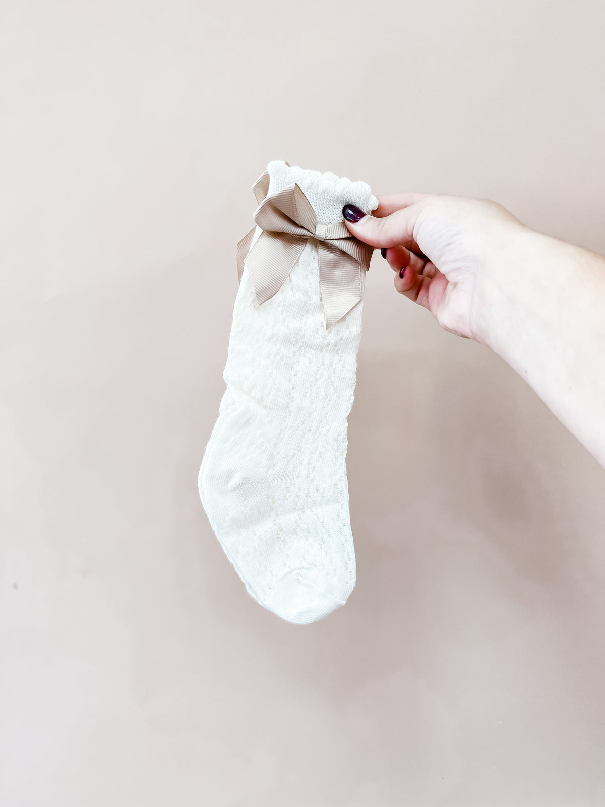 A hand with dark nail polish holds up a single Heirloom Bow Knee Sock by Tiny Tot Bundles, featuring a textured pattern and a large beige bow, against a plain light beige background.