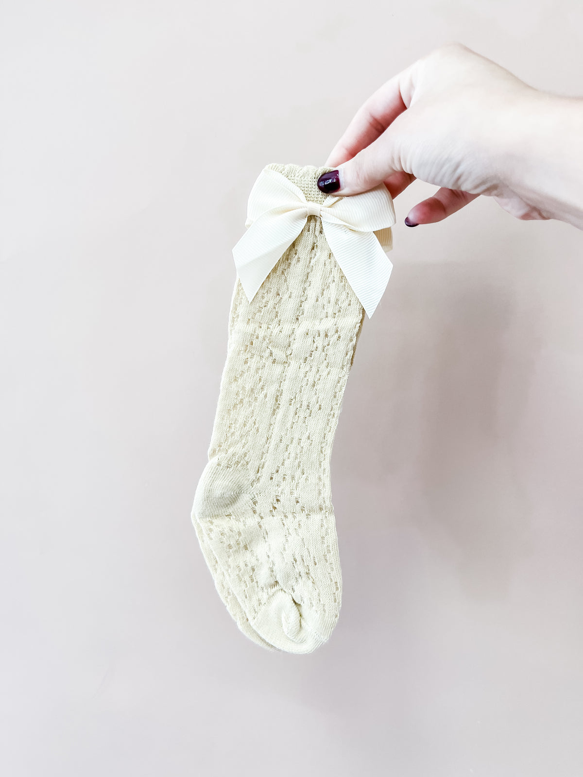 A hand with dark nail polish holds the Tiny Tot Bundles Heirloom Bow Knee Socks in pale yellow, featuring a large matching bow near the top, against a light beige background.