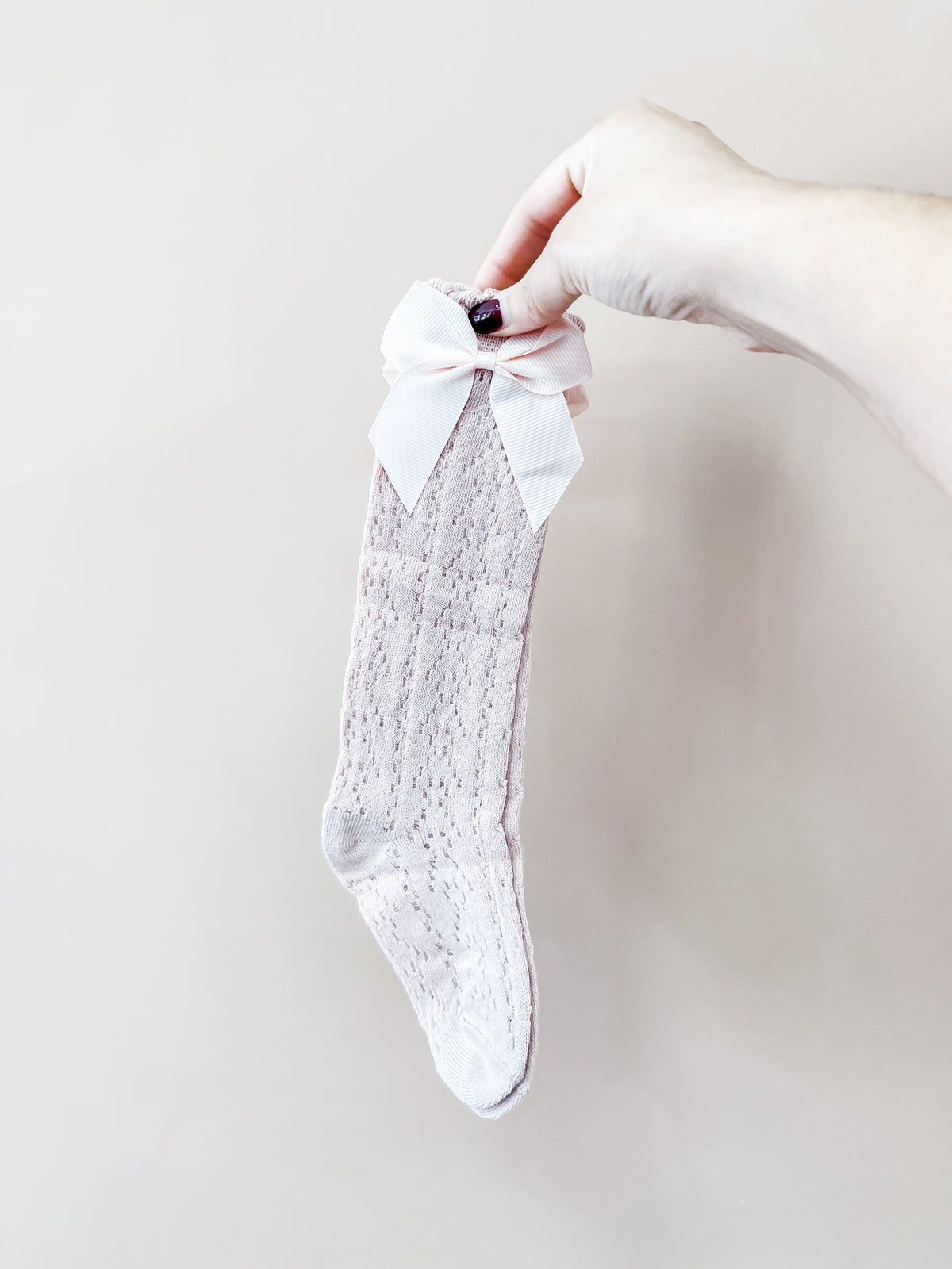 A hand holds a single Heirloom Bow Knee Sock by Tiny Tot Bundles, featuring a pale pink textured pattern and a large white ribbon bow at the top, set against a plain light background.
