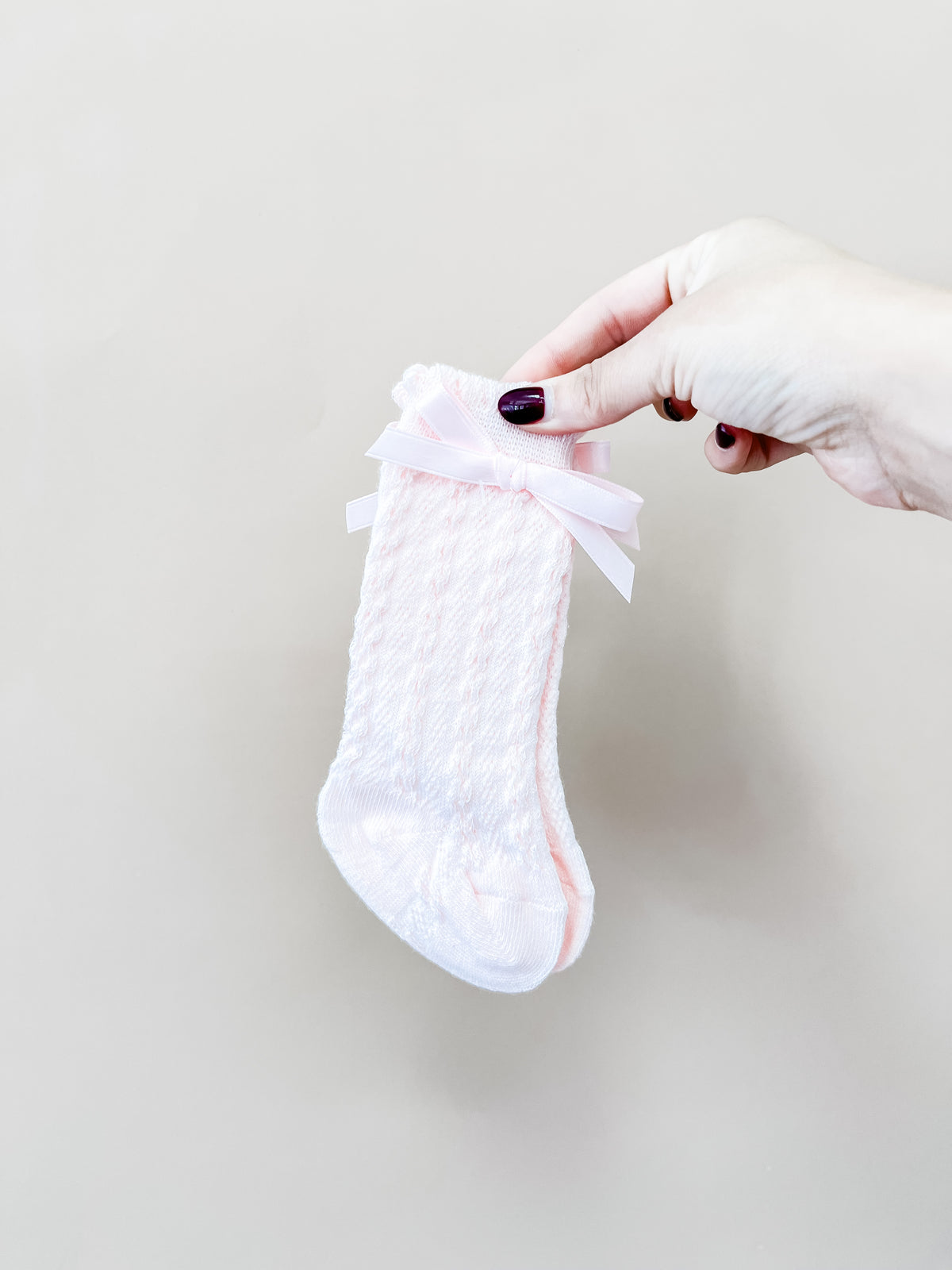 A hand with dark nail polish holds a pair of Tiny Tot Bundles Darlin Bow Knee Socks—white baby socks adorned with pink bows—against a plain beige background.