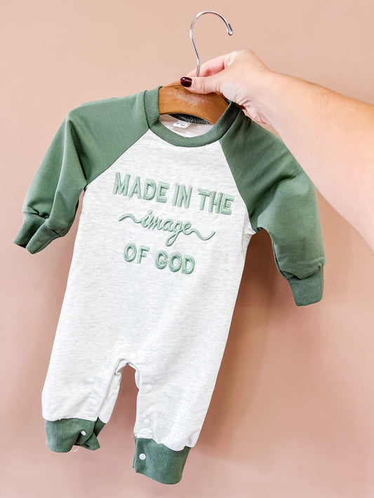 A hand holds the Tiny Tot Bundles Made In The Image Romper on a hanger. The green and white romper features “MADE IN THE IMAGE OF GOD” embroidered on the front, set against a solid light pink background.