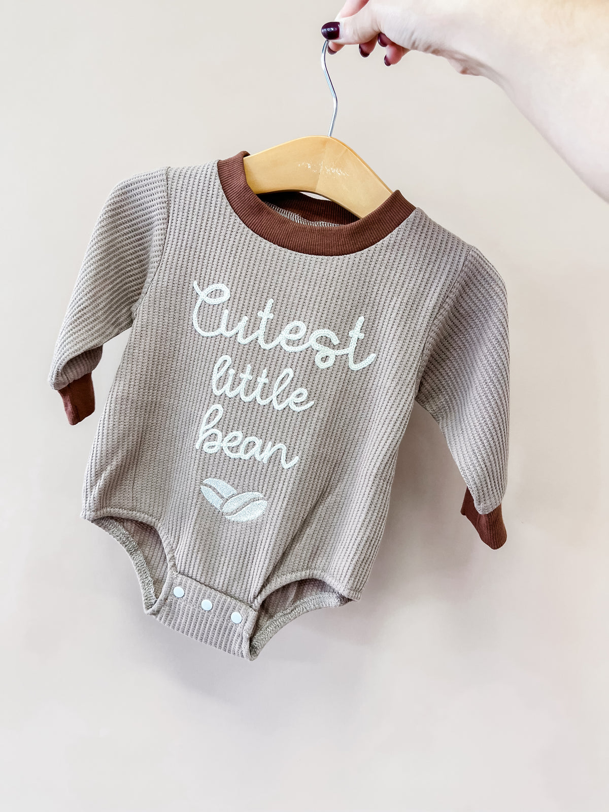 A hand holds a Tiny Tot Bundles Cutest Little Bean Romper on a hanger. The beige and brown romper features “Cutest little bean” and two small bean graphics printed on the front.