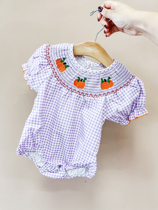 A hand holds up the Lilac Pumpkin Smocked Bubble by Tiny Tot Bundles—a purple and white gingham baby romper with orange pumpkin embroidery on the smocked collar.