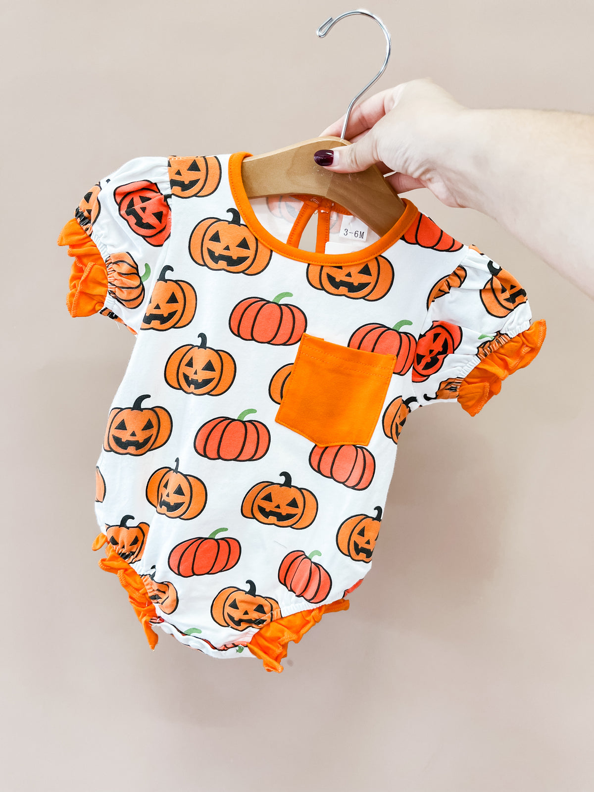 A hand holds the Pumpkin Patch Pocket Bubble by Tiny Tot Bundles, a baby romper with orange pumpkins, jack-o-lanterns, orange ruffled sleeves, a front pocket, and orange trim.