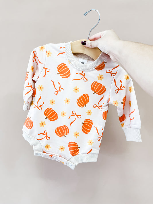 A hand holds the Sweet Pumpkin Bow Romper by Tiny Tot Bundles, featuring orange pumpkins, flowers, and bows on white fabric. The romper hangs on a plain, light-colored backdrop.