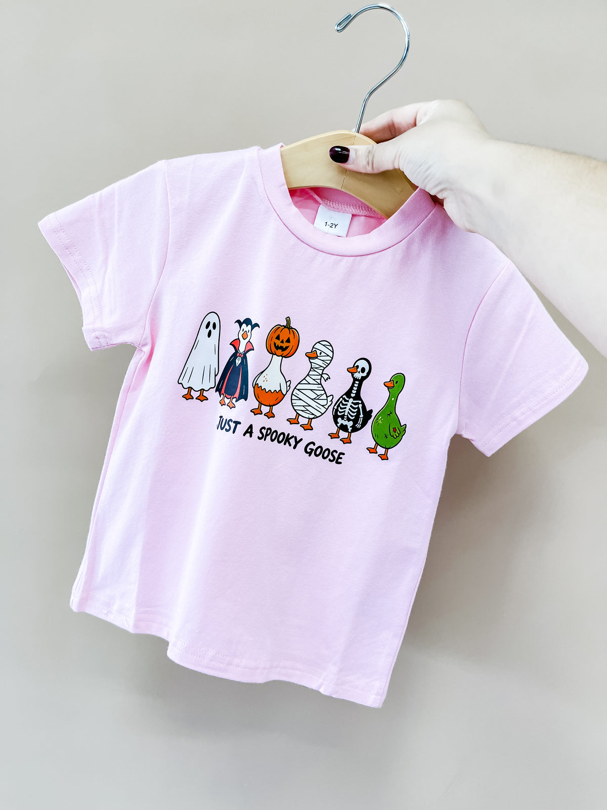 A hand holds the Pink Spooky Goose Tee by Tiny Tot Bundles, featuring cartoon geese in Halloween costumes and JUST A SPOOKY GOOSE text below the illustrations.