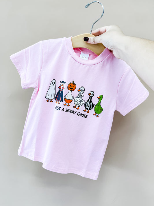 A hand holds the Pink Spooky Goose Tee by Tiny Tot Bundles, featuring cartoon geese in Halloween costumes and JUST A SPOOKY GOOSE text below the illustrations.
