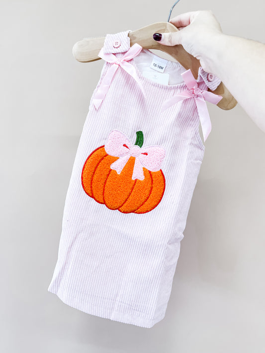 A hand holds the Sweet Pumpkin Corduroy Dress by Tiny Tot Bundles—a pink textured dress with an orange pumpkin and pink bow on the front, plus pink ribbon bows on the shoulder straps.