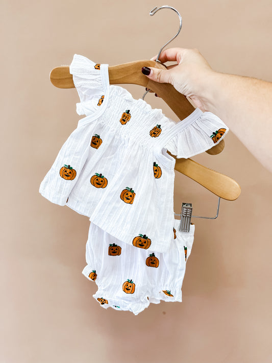 A hand holds the Pumpkin Peekaboo Set by Tiny Tot Bundles: a white baby outfit with orange pumpkin prints, featuring a ruffled top and matching bloomers, against a tan background.