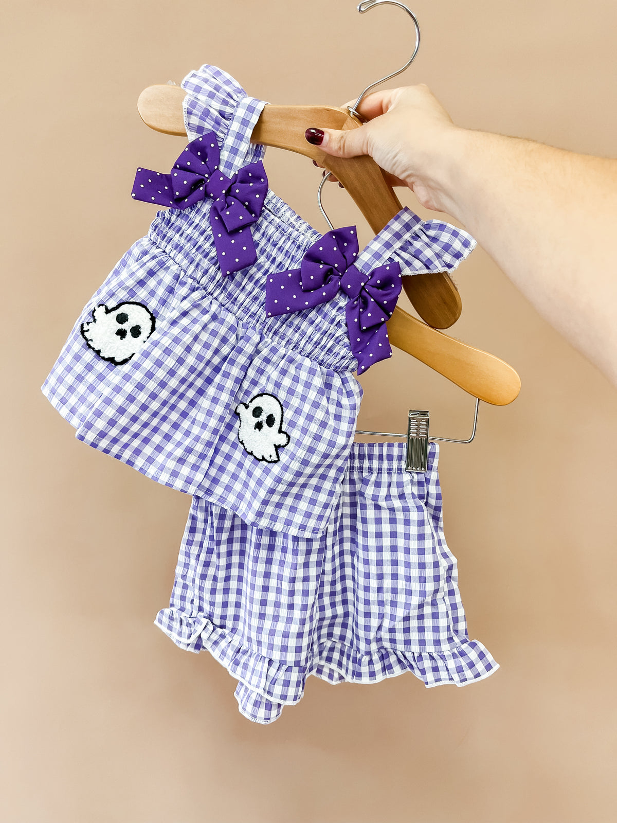 A hand holds the Purple Boo Ruffle Set by Tiny Tot Bundles, featuring purple gingham fabric, ruffles, bows, and ghost patches on wooden hangers against a beige background.