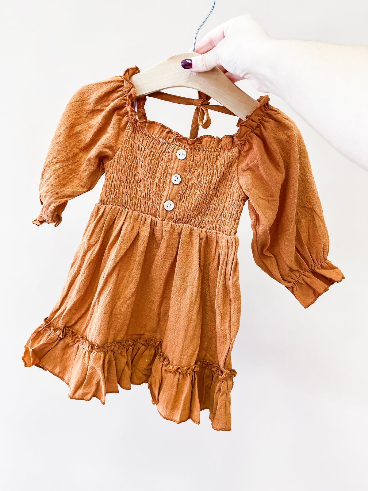 A hand holds a wooden hanger with the Rust & Ruffles Dress by Tiny Tot Bundles—a small, rust-colored long-sleeve dress featuring buttons, a ruffled hem, and gathered bodice against a plain light background.