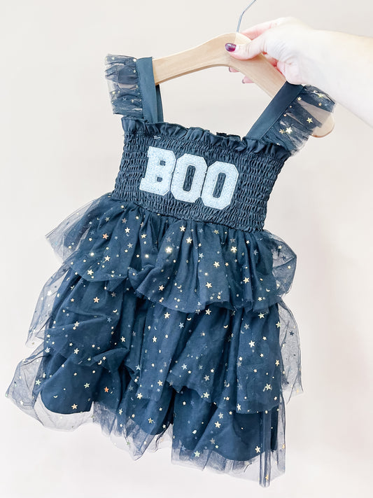 A hand holds up the Spooky Sparkle Tulle Dress by Tiny Tot Bundles, a child’s dark blue dress with BOO in white on the bodice and sparkly star patterns on the tulle ruffles.