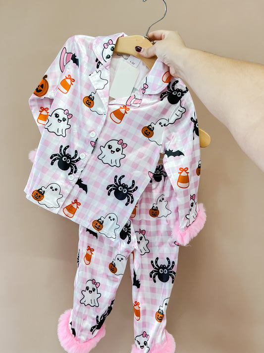 A hand holds up the Ghostly Glam PJ Set by Tiny Tot Bundles—a pink plaid pajama set with ghosts, bats, spiders, pumpkins, candy corn, and fuzzy pink cuffs on the sleeves and pants.