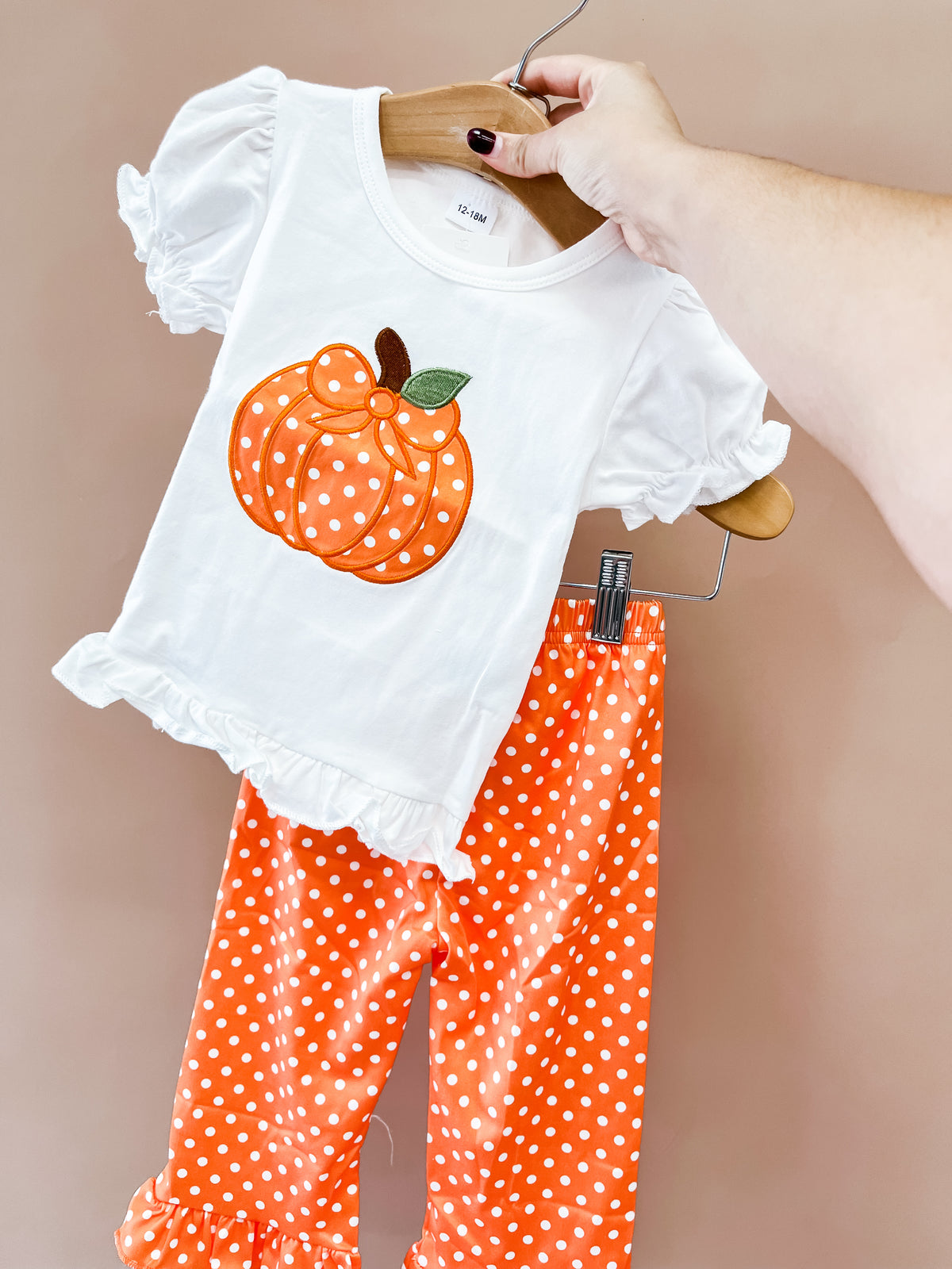 A hand holds the Tiny Tot Bundles Pumpkin Dot Ruffle Pant Set: a white shirt with a polka-dotted pumpkin and matching orange polka-dot ruffle pants, against a beige background.