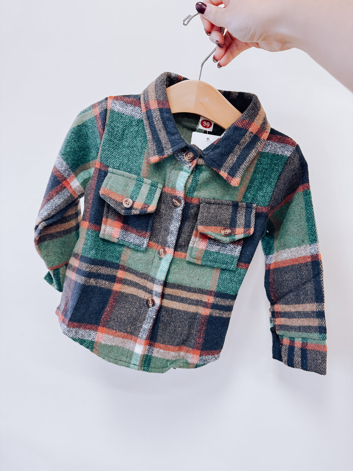 A hand holds a green, navy, and orange Evergreen Plaid Flannel shirt on a wooden hanger.