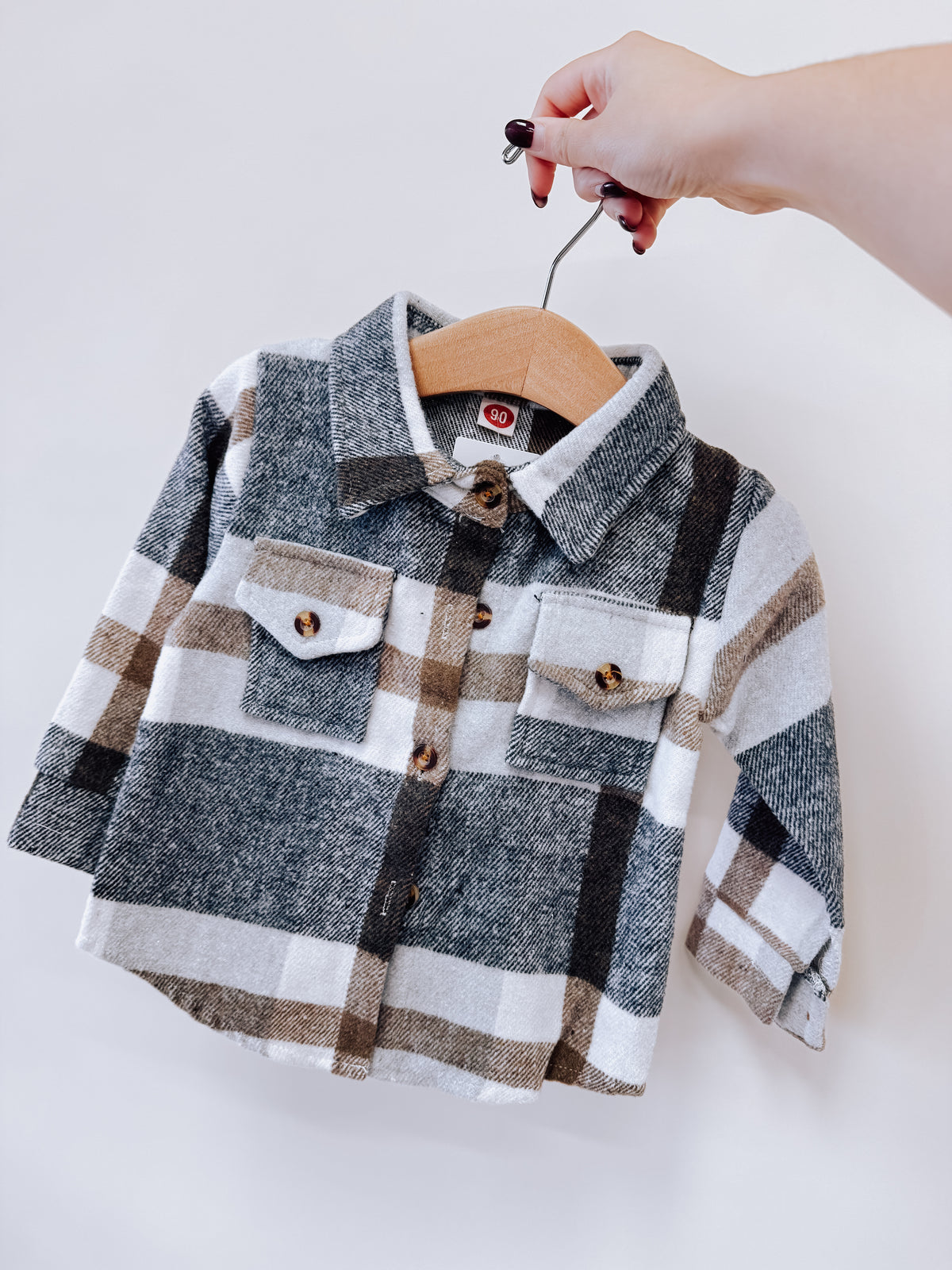 A hand holds a toddler-sized Neutrals Plaid Flannel featuring a grey, tan, and white plaid pattern with two chest pockets.