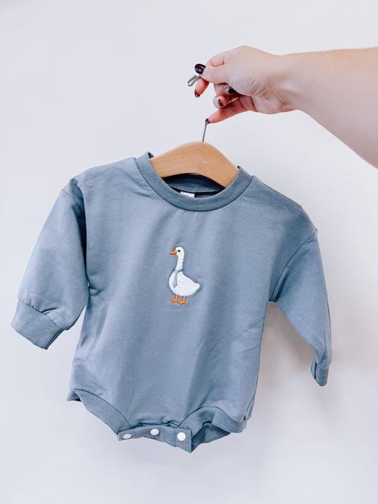 A hand holds a dusty blue Blue Goose Embroidered Bubble romper featuring an embroidered white goose on the chest.