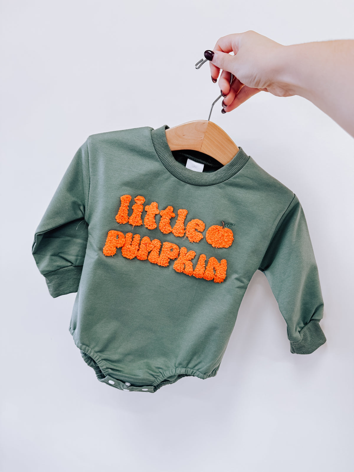 A hand holds a sage green long-sleeved Tiny Tot Bundles Little Pumpkin Chenille Romper on a hanger, featuring the orange fuzzy text little pumpkin with a pumpkin icon on the chest.