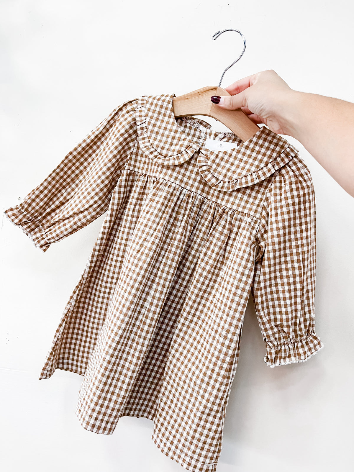 A hand holds a Tiny Tot Bundles Autumn Brown Collar Dress, a brown and white gingham long-sleeve dress with a rounded Peter Pan collar, on a wooden hanger.