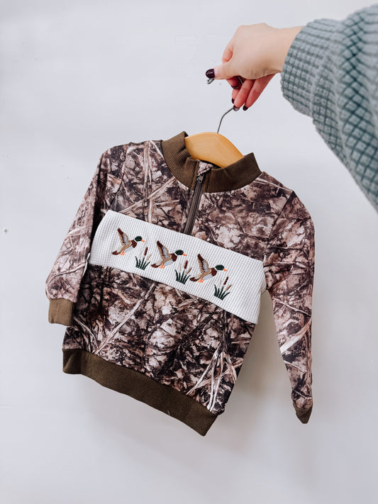A Tiny Tot Bundles Camo Smocked Duck Pullover featuring a camo-print with a white smocked chest panel and embroidered mallard ducks, displayed on a wooden hanger.