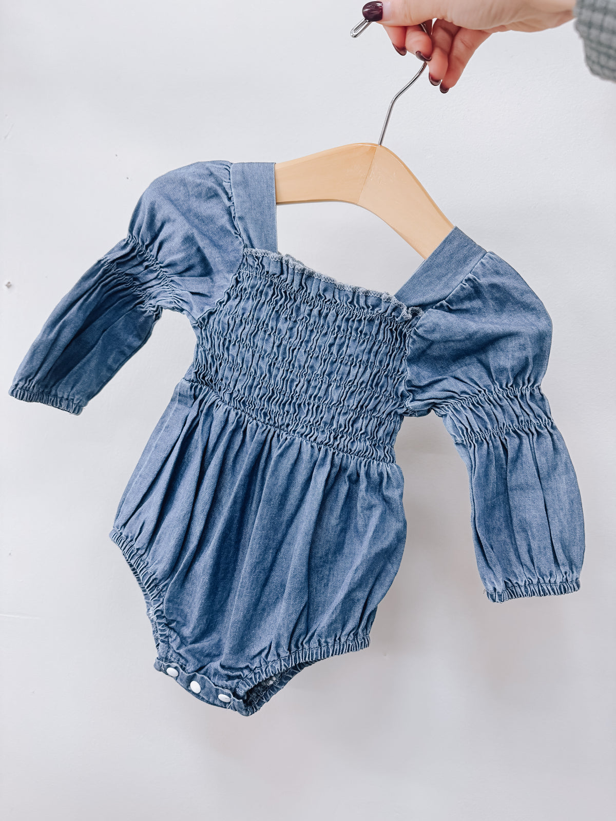 Tiny Tot Bundles Denim Blue Romper featuring a long-sleeved blue denim design with a smocked bodice and bottom snaps, displayed on a wooden hanger.