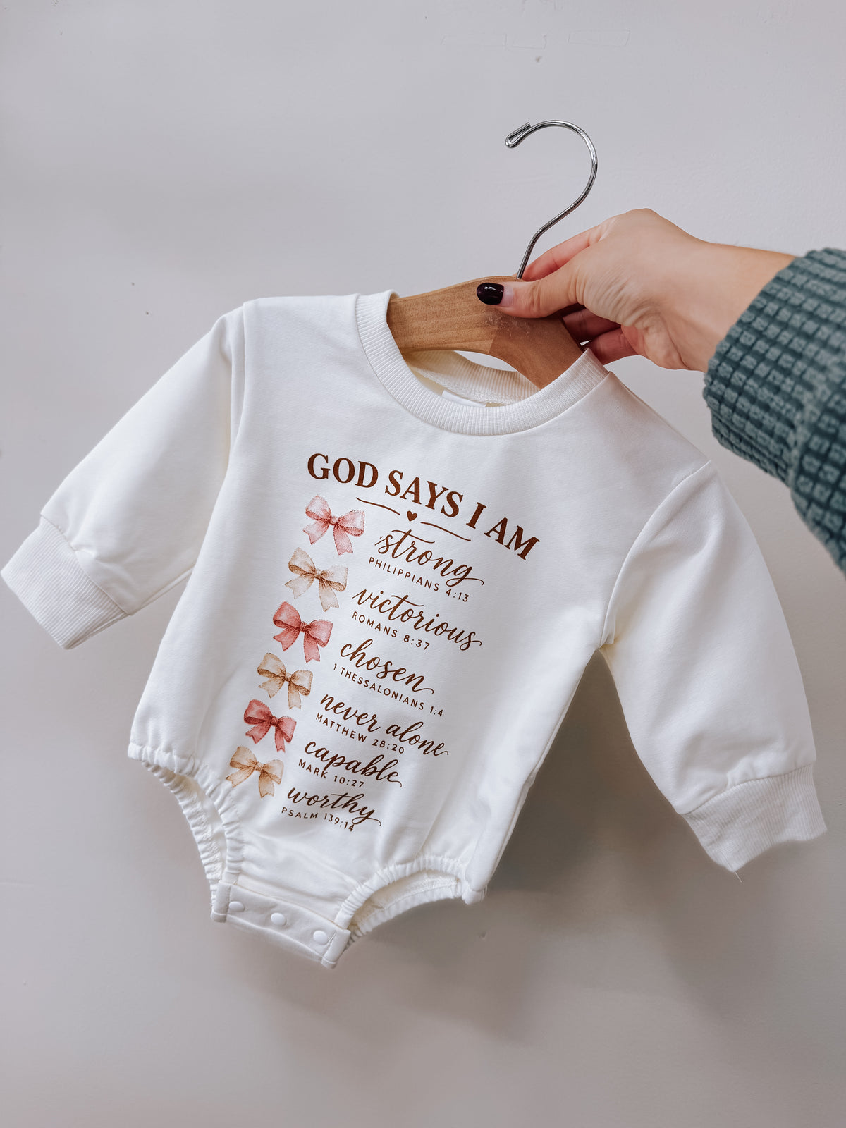 A white long-sleeved God Say’s I Am Pink Bubble bodysuit printed with six bows and the text God says I am strong, victorious, chosen, never alone, capable, worthy, each followed by a Bible verse.