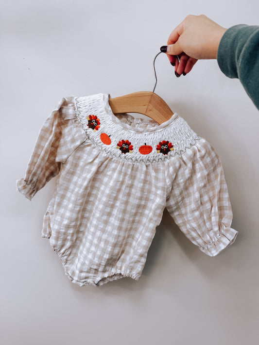 Tiny Tot Bundles Smocked Turkey Romper: a long-sleeved beige gingham romper with a smocked neckline embroidered with small turkeys and pumpkins.
