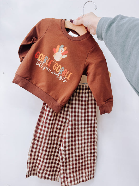 Tiny Tot Bundles Gobble Gobble Plaid Two-Piece Set consisting of a brown long-sleeve sweatshirt with a turkey graphic and the text Gobble Gobble till you wobble, paired with brown and white plaid pants.