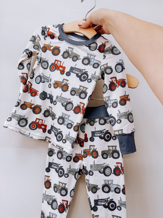 A Tiny Tot Bundles Tractor Trails Two Piece Set featuring a long-sleeve shirt and matching pants with a repeating tractor print and dark gray trim, displayed on a wooden hanger.