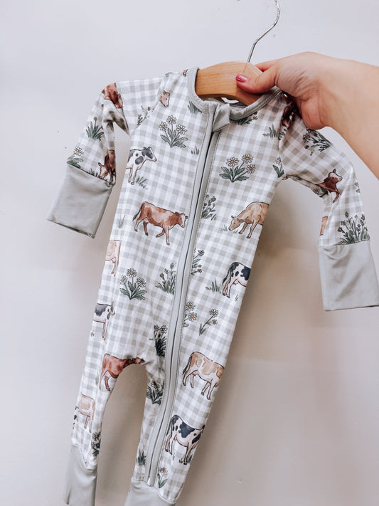 Tiny Tot Bundles Pasture & Plaid Zippy long-sleeved infant footed sleeper with a zipper closure, featuring a pattern of cows and wildflowers on a grey gingham background.