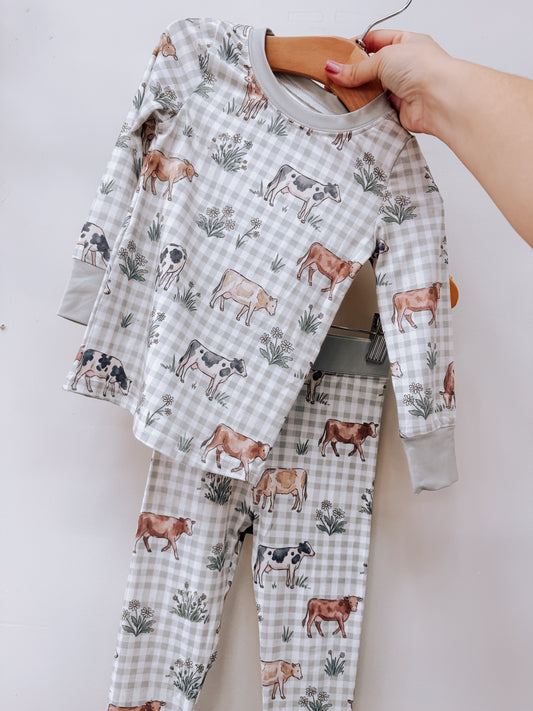 Pasture & Plaid Two Piece Set featuring a grey gingham long-sleeved shirt and pants with illustrations of cows and small flowers.