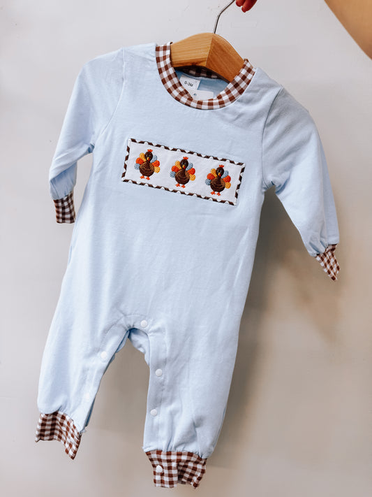Tiny Tot Bundles Smocked Turkey Gingham Romper in light blue, featuring a central panel with three embroidered turkeys and brown gingham trim on the neckline, cuffs, and leg openings.