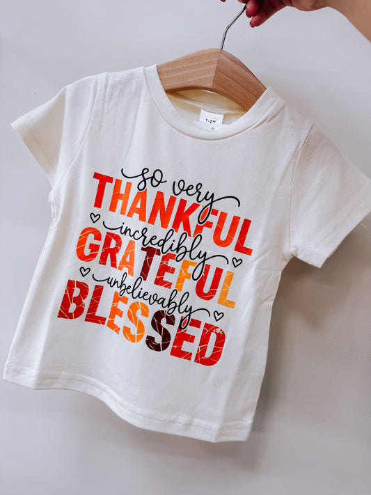Tiny Tot Bundles Thankful Grateful Blessed Tee on a wooden hanger, featuring cream-colored fabric with the text so very THANKFUL incredibly GRATEFUL unbelievably BLESSED in red, orange, and brown lettering.