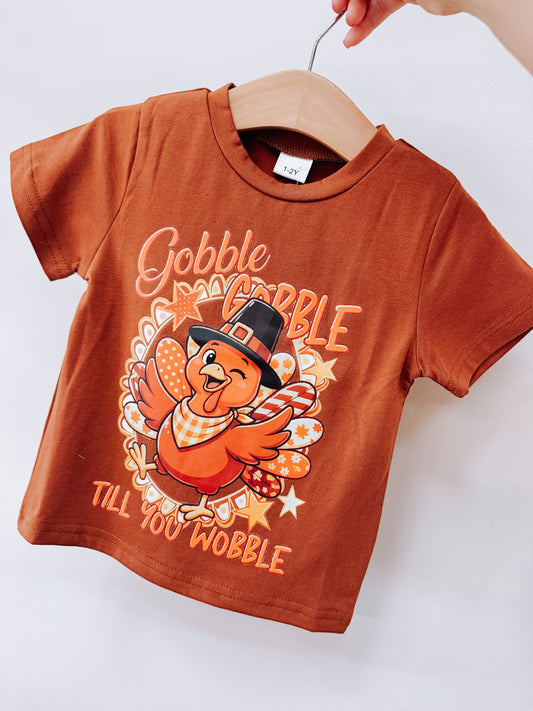 Rust-colored Tiny Tot Bundles Gobble Till You Wobble Tee featuring a graphic of a turkey wearing a pilgrim hat, surrounded by stars and the text Gobble Gobble Till You Wobble.