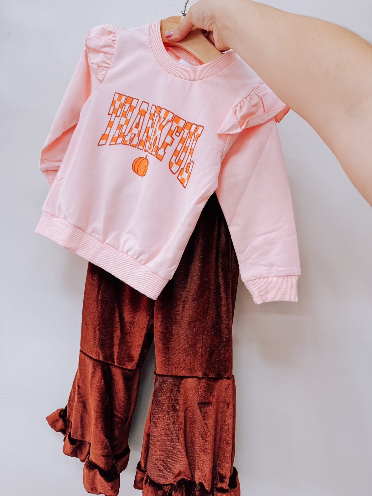 A hand holds up a pink long-sleeved sweatshirt with the word THANKFUL and a small pumpkin graphic, paired with dark brown velvet bell-bottom pants from the Thankful Ruffle Bell Bottom Set by Tiny Tot Bundles.